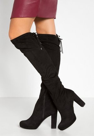 Femme portant des bottes hautes en daim noir à talons, associées à une courte jupe bordeaux, debout jambes croisées sur fond blanc.
