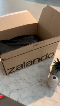 Caixa de cartão castanha, rotulada "zalando", parcialmente aberta, revelando uma peça de vestuário preta no interior. Superfície de mármore circundante, pequeno vaso branco e planta.