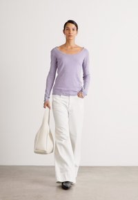Lys lilla langermet topp kombinert med hvite vide bukser. Modellen bærer en beige skulderveske og har på seg svarte sko. Enkelt bakgrunn.