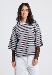 Young woman with wavy hair wearing a loose striped t-shirt over a long-sleeve lace top and light gray sweatpants, standing against a plain background.