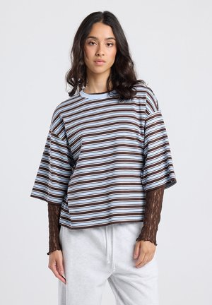 Jeune femme avec des cheveux ondulés portant un t-shirt à rayures loose sur un haut en dentelle à manches longues et un pantalon de survêtement gris clair, se tenant contre un fond uni.