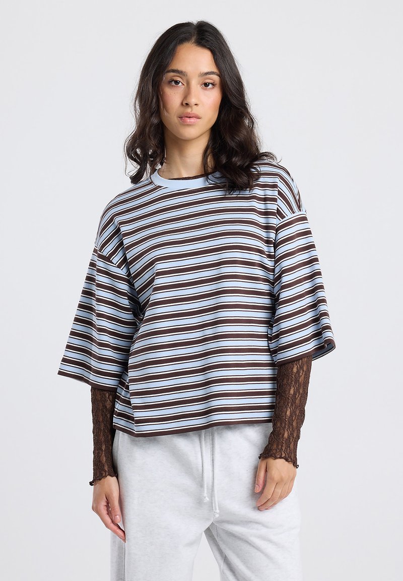 Young woman with wavy hair wearing a loose striped t-shirt over a long-sleeve lace top and light gray sweatpants, standing against a plain background.