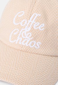 Căciulă dintr-un model herringbone în piersiciu deschis, cu text alb brodat "Cafea & Haos." Este dotată cu o vizieră curbată și un material texturat.