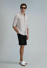 Chemise en lin beige clair avec manches retroussées, short noir et baskets blanches avec semelles marron ; modèle portant des lunettes de soleil noires.