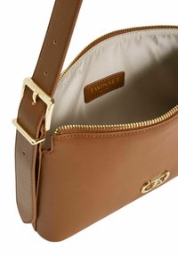 Borsa in pelle tan con superficie liscia, accessori in oro e chiusura a zip. L'interno presenta una fodera crema e un'etichetta del marchio.