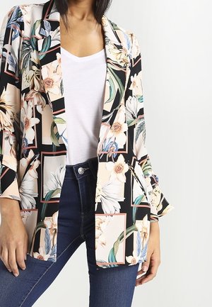 Kvinde iført blomsterprintet blazer med hvide, grønne og beige blomster, hvid t-shirt og mørkeblå jeans, stående mod en ensfarvet baggrund.