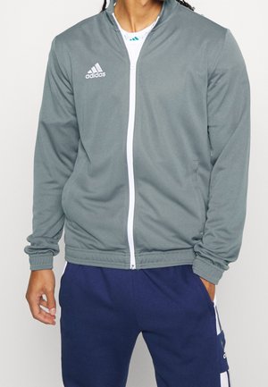 Persoon draagt een grijze Adidas jas met ritssluiting en witte bies, over een wit shirt, en donkerblauwe Adidas trainingsbroek.