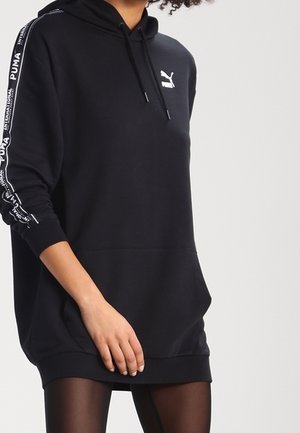 Frau trägt ein schwarzes Puma-Kapuzenkleid mit Logo auf der Brust und markanten Streifen an den Ärmeln, kombiniert mit schwarzen durchsichtigen Strumpfhosen.