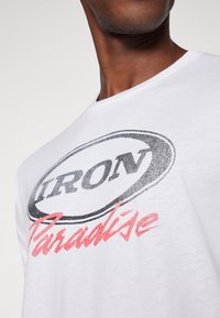 Balta medvilninė marškinė su ryškiu juodu užrašu "IRON" ir raudonu užrašu "Paradise" paryškintu šriftu. Apvalus iškirpimas ir trumpomis rankovėmis.