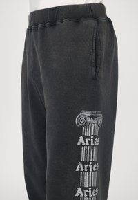 Schwarze Baumwoll-Sweatpants mit Kordelzugbund, Seitentaschen und einem bedruckten Säulendesign mit dem Text "Aries" auf dem linken Bein.