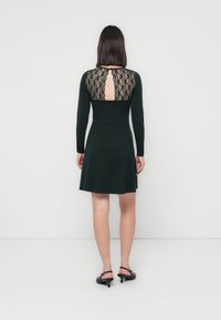 Robe noire en dentelle au dos, avec des manches longues, un corsage ajusté et une jupe évasée. Tissu lisse, longueur genou, avec une découpe en forme de clé dans le dos.