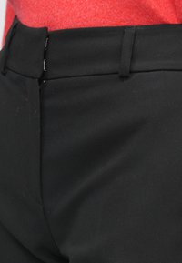 Melnas bikses ar gludu tekstūru, plakanu jostasvietu ar mazām metāla āķīšiem un redzamu rāvējslēdzēju, daļēji redzamas uz sarkana fona.