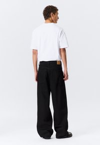 Pantalons larges noirs avec poches avant et arrière, portés avec un t-shirt blanc uni. Tissu en coton avec une texture lisse et étiquette en cuir marron.
