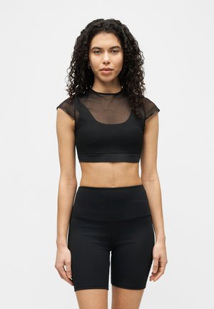 Femeie cu păr creț, purtând un top negru transparent, scurt, peste un sutien sport negru și pantaloni scurți negri de biciclist, cu talie înaltă, stând în fața unui fundal simplu.