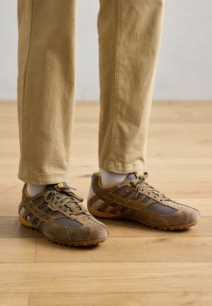 Zapatillas deportivas con un upper de ante y malla gris, acentos amarillos, suela de goma texturizada y diseño de cordones. Los pantalones son caquis.