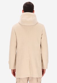 Uomo con cappotto in corduroy a coste beige e colletto alto, visto da dietro, in piedi contro uno sfondo chiaro e uniforme.
