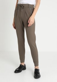 Bruine taps toelopende broek met een tailleband met trekkoord, voorzien van ritssluitingen en een gladde textuur, gecombineerd met zwarte veterschoenen.