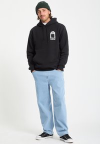 Haut à capuche noir avec un design graphique sur la poitrine, associé à un jean bleu clair et des baskets noires. Le modèle porte un bonnet vert.
