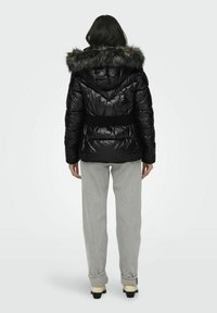 Chaqueta acolchada negra con capucha de pelo sintético, acabado metálico, cintura con cinturón y textura acolchada. La modelo lleva pantalones y botas gris claro.