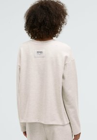 Light beige sweatshirt with a relaxed fit, ribbed neckline, and a rectangular label featuring "SENSES" on the upper back. Soft texture.