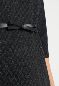 Svart klänning med ett texturerat mönster och en glänsande finish. Den har ett tunt svart band som är knutet i en rosett i midjan.