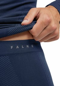 Hand pulling up navy blue textured waistband underwear with "FALKE" logo, showing part of bare skin and matching textured clothing sleeve.