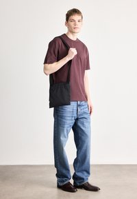 Črna torba za nošenje čez ramo, izdelana iz blaga. Model nosi bordo majico in modre kavbojke s temnimi usnjenimi čevlji. Nevtralno ozadje.