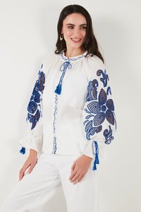 Femeie purtând o bluză albă cu broderie florală albastră pe mâneci bufante și pantaloni albi, zâmbind și uitându-se în lateral.