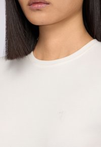 Světle béžové bavlněné tričko s kulatým výstřihem a krátkými rukávy. Na přední straně je decentně embosované logo. Hladká textura.