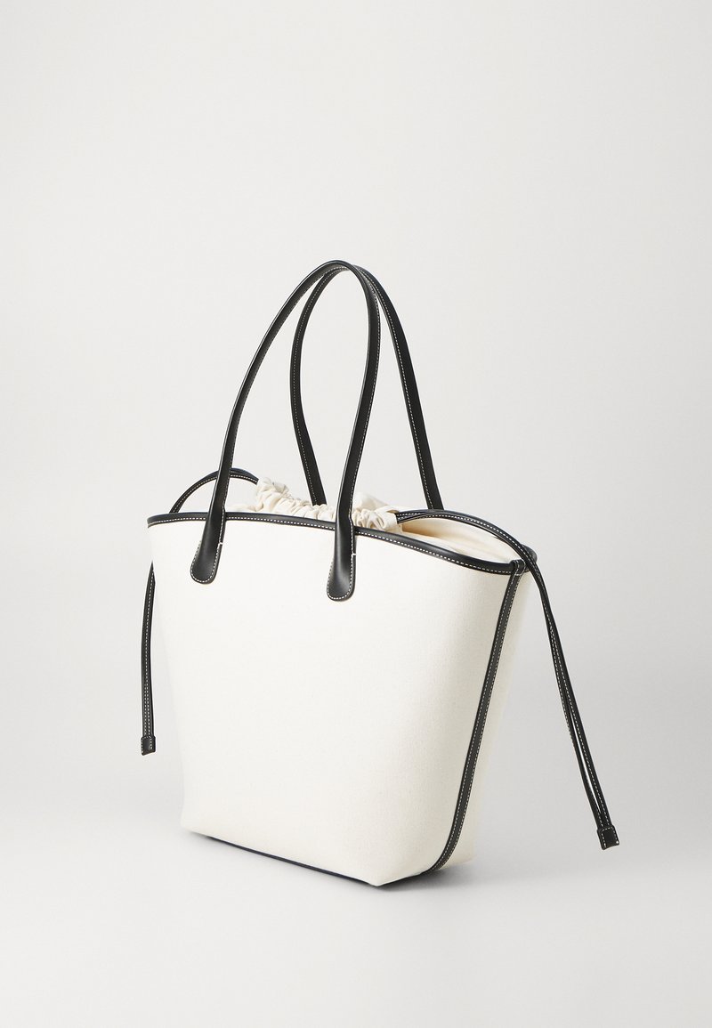 Tote bag en toile blanche avec des accents en simili cuir noir, équipé de deux poignées, d'une fermeture à cordon et de coutures blanches contrastantes.