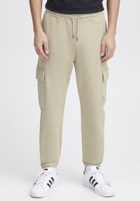 Khaki cargojoggers met elastische tailleband en boorden, twee zijzakken op de dijen en een gladde, gestructureerde stof. Gecombineerd met witte sneakers.