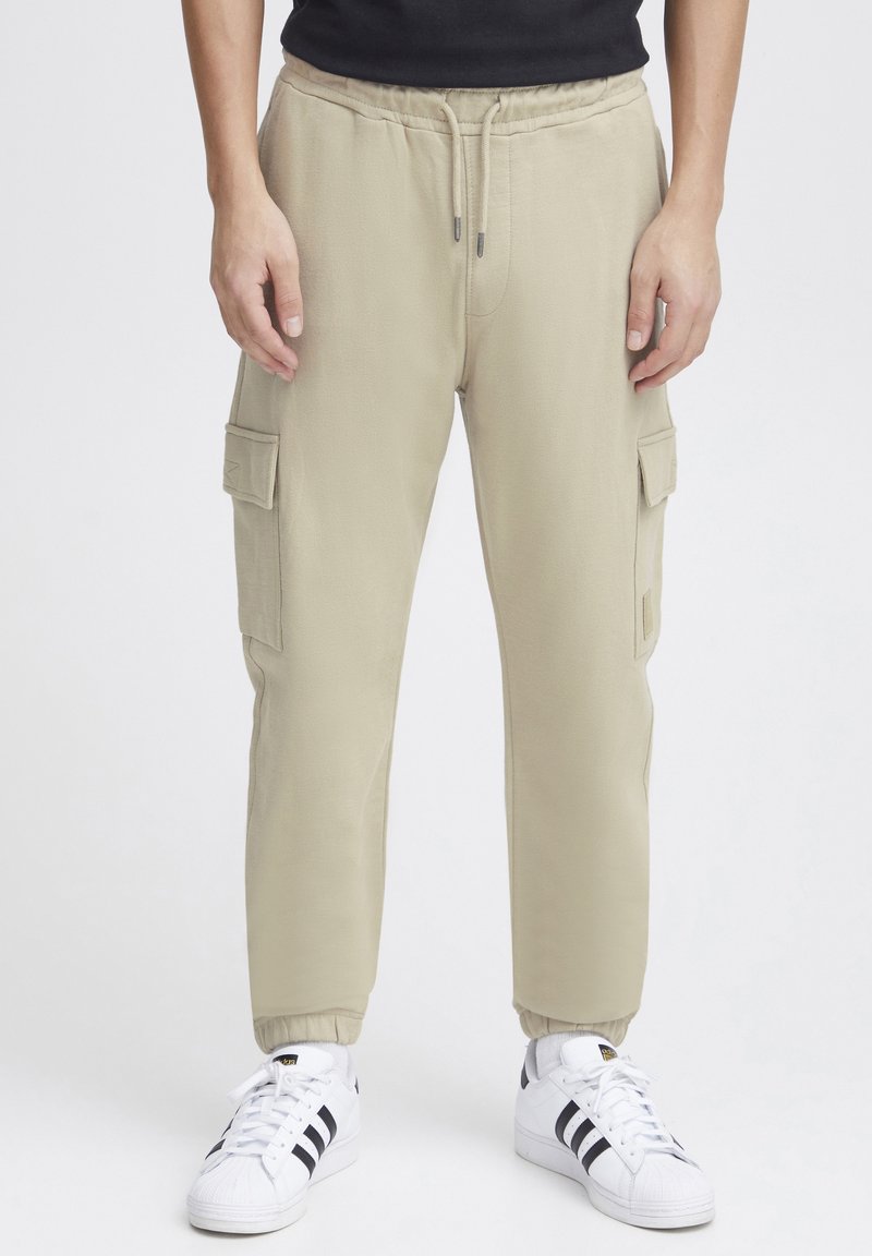 Khaki cargojoggers met elastische tailleband en boorden, twee zijzakken op de dijen en een gladde, gestructureerde stof. Gecombineerd met witte sneakers.