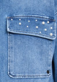 Tascone di giacca di jeans blu chiaro con decorativi stud in perla disposti sulla patta. Presenta cuciture e un bottone rotondo nero.