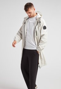 Giacca impermeabile grigio chiaro con cappuccio, polsini elastici e dettagli neri, abbinata a pantaloni neri e una t-shirt bianca sotto.