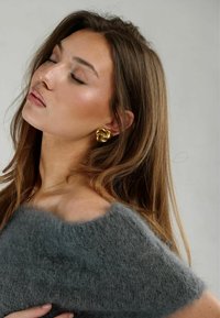Maglione grigio, morbido e lavorato a maglia con design off-shoulder. Il modello indossa un orecchino a forma di nodo dorato, la texture morbida è visibile nel maglione.