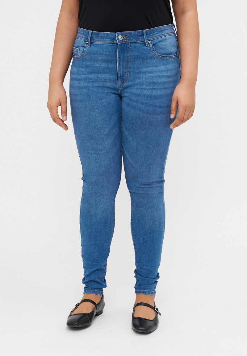 Skinny jeans in medium blue denim, featuring a high waist, five pockets, and subtle fading. Paired with black flat shoes.
