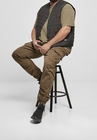 Vadderad svart väst över olivgrön t-shirt, kombinerad med olivgröna cargobyxor och svarta sneakers. Modellen sitter på en pall.