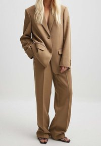 Blazer oversized marron avec deux boutons et revers à pointe, associé à un pantalon large assorti. Tissu lisse et texturé, avec des accents minimalistes.