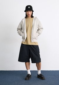 Lys grå zip-up hettegenser, beige t-skjorte, svarte shorts, hvite sokker, svart bøttehatt, og svarte sko med tykke såler, stående på et blått teppe.