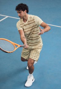 Mužský tenista v béžovém pruhovaném tričku a kostkovaných šortkách odpaluje forhent na modrém krytém kurtu s oranžovým raketou.