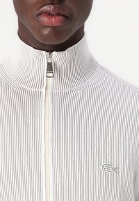 Biely rebrovaný sveter na zips s šedými pruhmi, vysokým golierom a vyšívaným logom Lacoste na ľavom hrudi, so strieborným zipsom.
