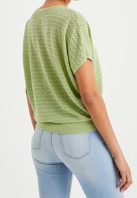 Vihreä lime neulepaita lyhyillä hihoilla, joka esittelee kiemuraista tekstuuria ja rentoa istuvuutta. Yhdistettynä vaaleansinisiin farkkuihin.