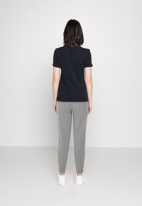 Person i svart kortärmad skjorta, grå träningsbyxor och vita sneakers som står vänd mot en enkel vit vägg.