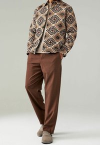 Giacca con motivo geometrico nei colori marrone, beige e nero, abbinata a pantaloni marroni. Le calzature sono di un grigio chiaro con un design semplice.