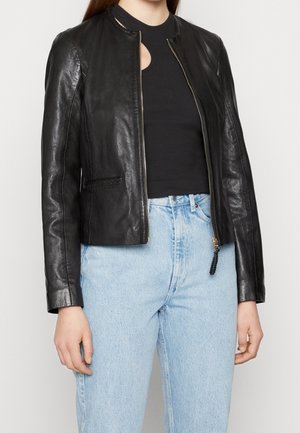 Femme portant une veste en cuir noire sur un haut noir et un jean taille haute bleu clair, debout devant un fond clair uni.