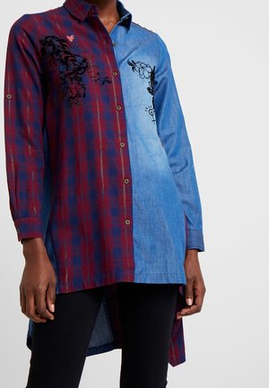 Woman wearing a long-sleeve, button-up shirt with one half red plaid and the other half light blue denim with floral embroidery.