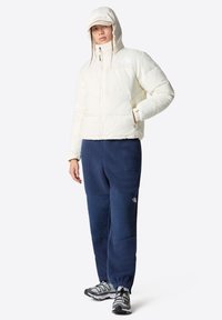 Giacca puffer bianca con cappuccio, chiusura con zip e logo. Abbinata a pantaloni in poliestere blu navy con texture. Scarpe da trekking nere e grigie.