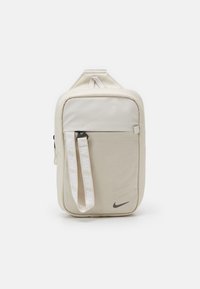 Beige Nike crossbody-väska med framficka, märkt vit rem och svart Nike-logotyp i nedre högra hörnet.
