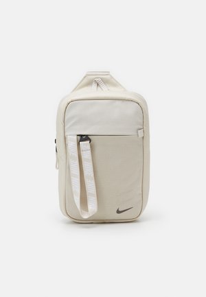 Borsa a tracolla beige Nike con tasca anteriore, tracolla bianca con logo e marchio, e logo Nike nero nell'angolo in basso a destra.