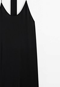 Robe noire sans manches à fines bretelles suspendue devant un mur blanc.
